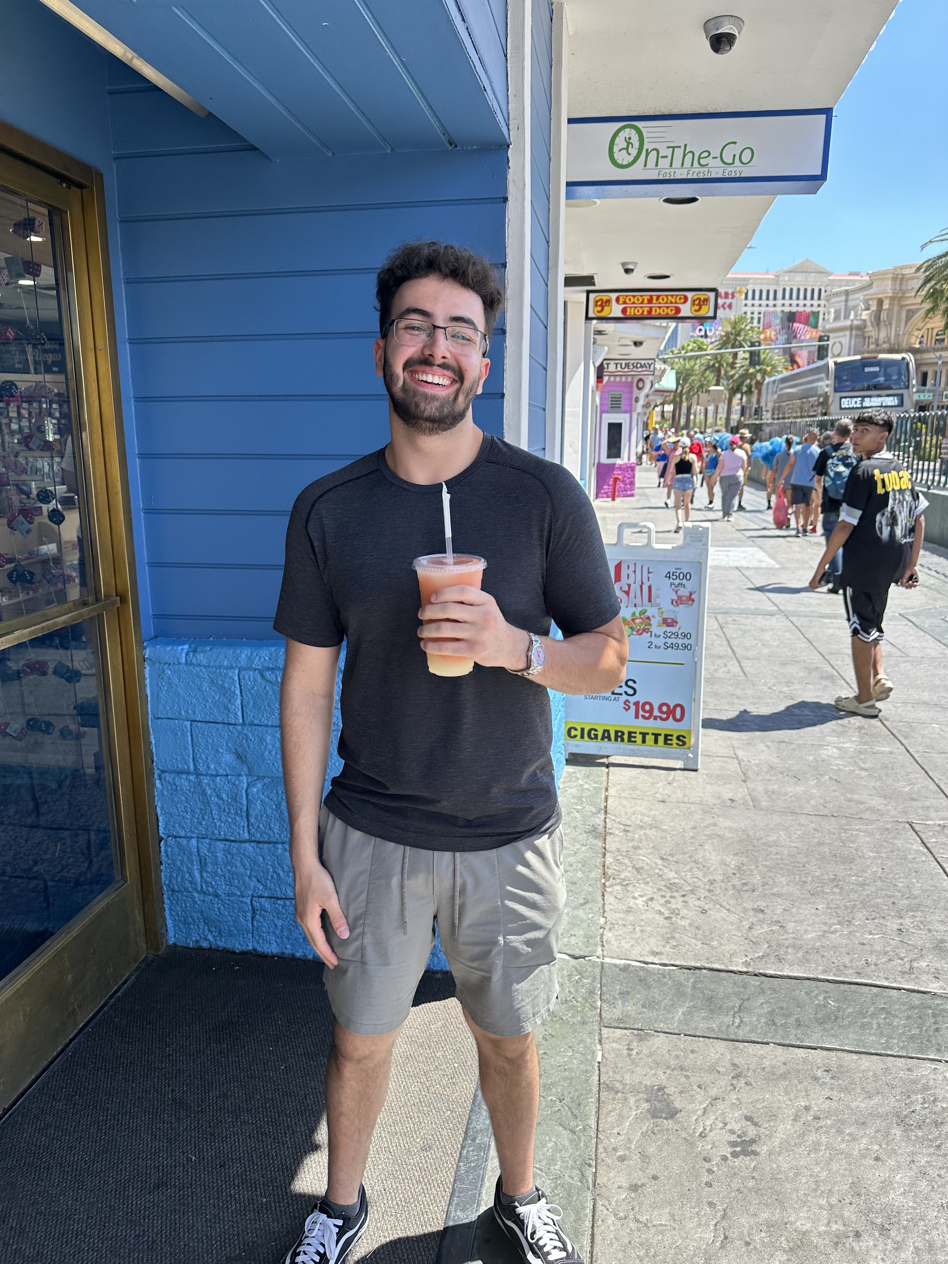 Profile picture of Karim Maftoun, standing outdoors on a sunny day,  holding a beverage in a clear cup with a straw. They are wearing a dark t-shirt and light-colored shorts, and are positioned in front of a blue building. The background includes a sidewalk with a advertising, as well as a few people walking in the distance.